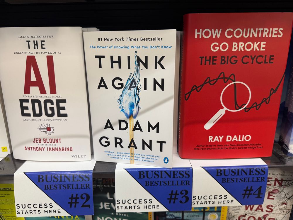 A shelf of “business bestsellers” at a store inside Boston Logan airport. The titles in sequence read “The AI Edge,” “Think Again,” “How Countries Go Broke”