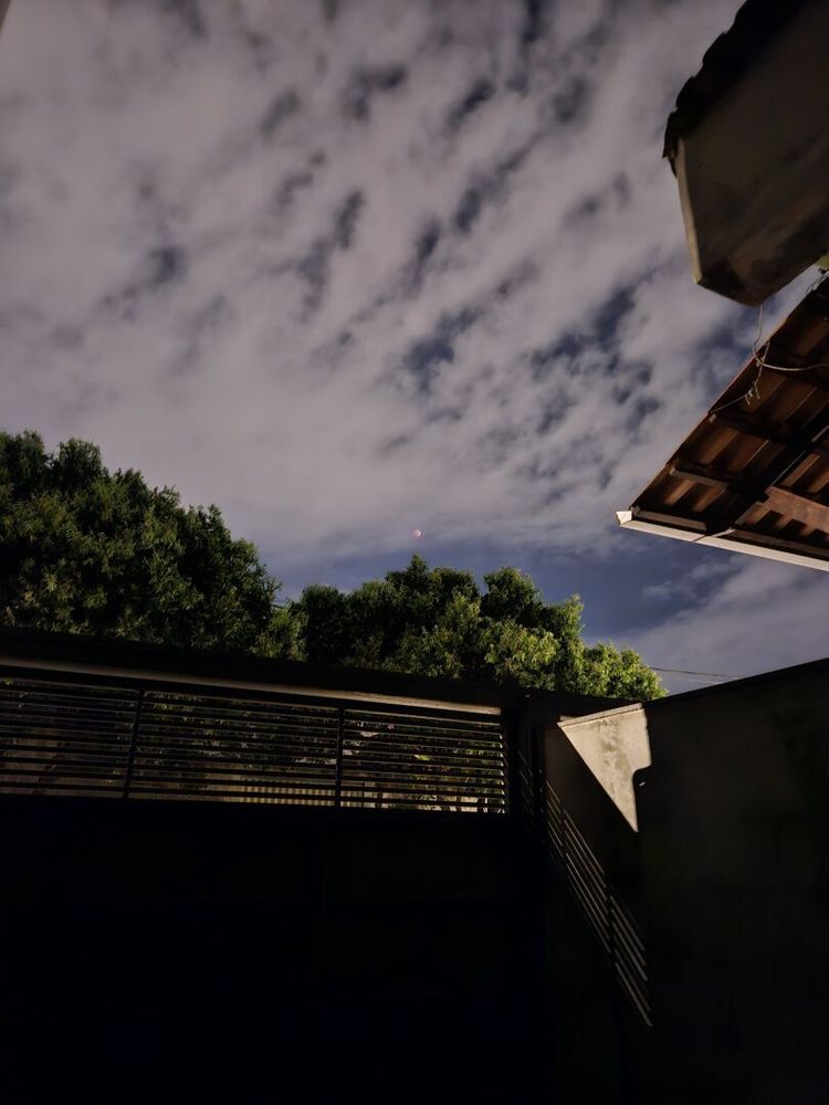 A nighttime scene with a cloudy sky. A reddish, partially obscured Blood Moon is faintly visible through the clouds. The foreground includes dark silhouettes of rooftops, a fence, and treetops. The primary and nearest light source is a street light pole (not directly visible in the image), casting a glow on the surrounding area. The overall lighting is low.