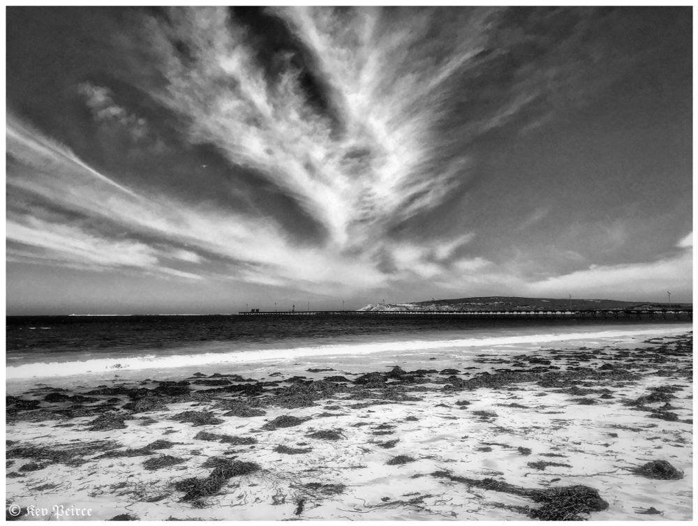 Black and white coastal scene with seaweed strewn across a quiet beach. Gentle waves lap the shore beneath a dramatic sky filled with wispy, textured clouds.

A long pier stretches into the distance, pointing toward a low hill or island on the horizon. The image is signed “Kev Peirce” in the bottom left corner.
