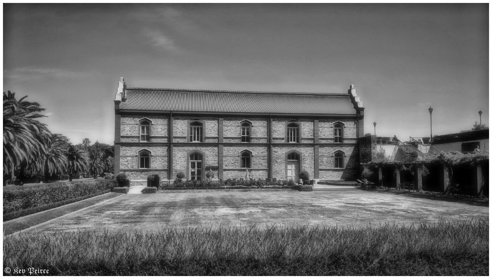 Black and white photo of a historic stone building with arched windows, surrounded by palm trees and manicured gardens.

A grassy lawn stretches across the foreground. On the right, a shaded walkway with benches adds a calm, contemplative touch. The monochrome palette highlights texture and timelessness.