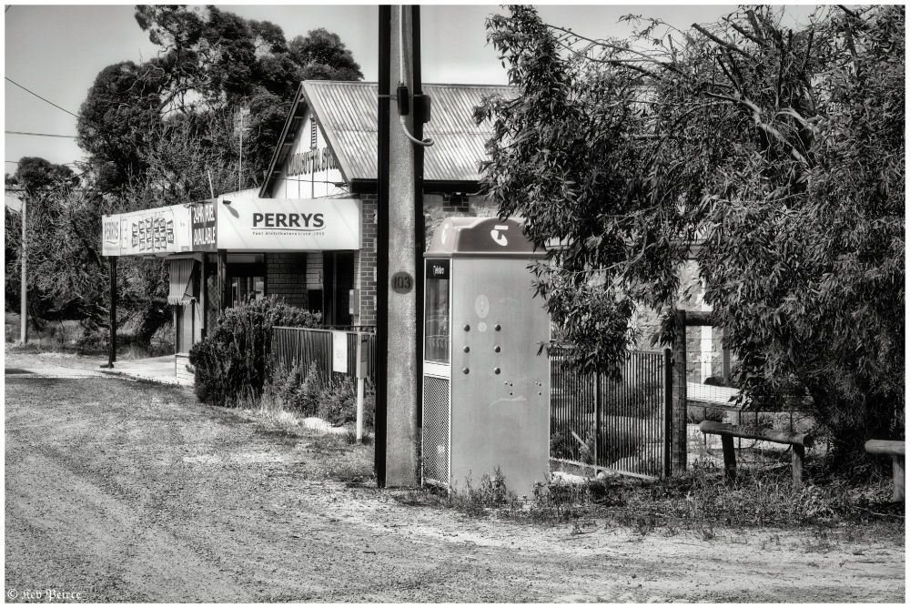 Black-and-white photo of Perrys store in Kyancutta, South Australia.

The foreground features a dirt road and an old-style telephone booth. The store has a corrugated metal roof, trees flanking both sides, and a vintage sign reading ‘PERRYS Fuel distributor since 1949’.

The rural setting and monochrome palette suggest a quiet, timeless atmosphere.