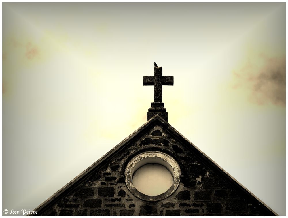 A single bird rests on the peak of a stone cross mounted above a circular window on a weathered rooftop.

The soft gradient of the sky—yellow bleeding into gray—casts a tranquil mood over the scene, evoking silence and reflection.