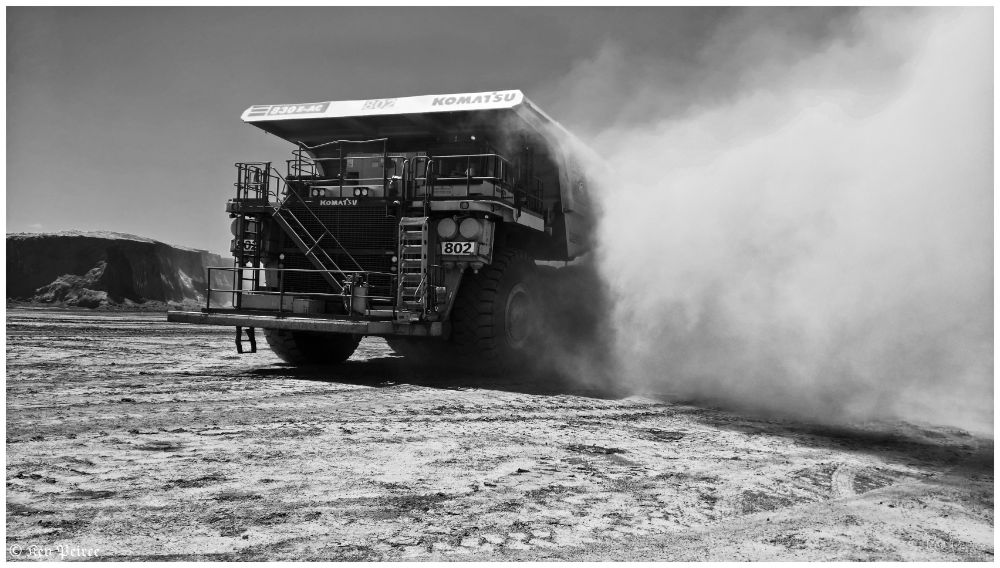 A black and white photograph of a Komatsu 830E-AC haul truck, numbered 802, rumbling through a rugged landscape.

The massive vehicle kicks up thick clouds of dust as it moves, its oversized wheels and heavy-duty frame silhouetted starkly against the swirling backdrop.

The grayscale tones emphasize the industrial power and raw environment, giving the image a dramatic, timeless feel.