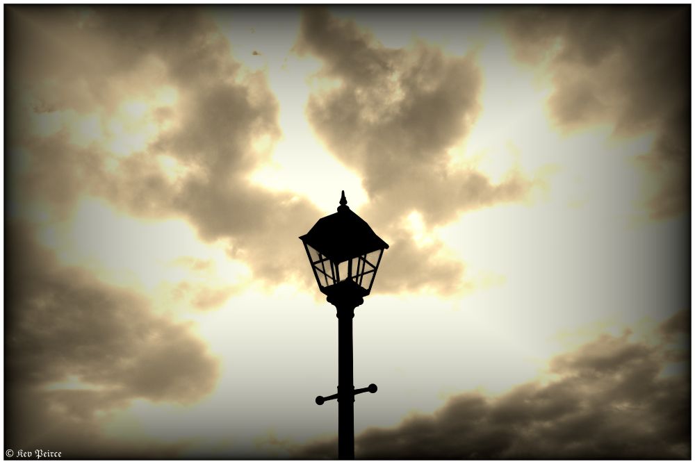 A tall, vintage lamp post with glass-paneled lantern stands silhouetted against a sepia-toned sky filled with thick, textured clouds.

The atmosphere is nostalgic and dramatic, with diffused sunlight filtering through the cloud cover, emphasizing the aged elegance of the lamp.