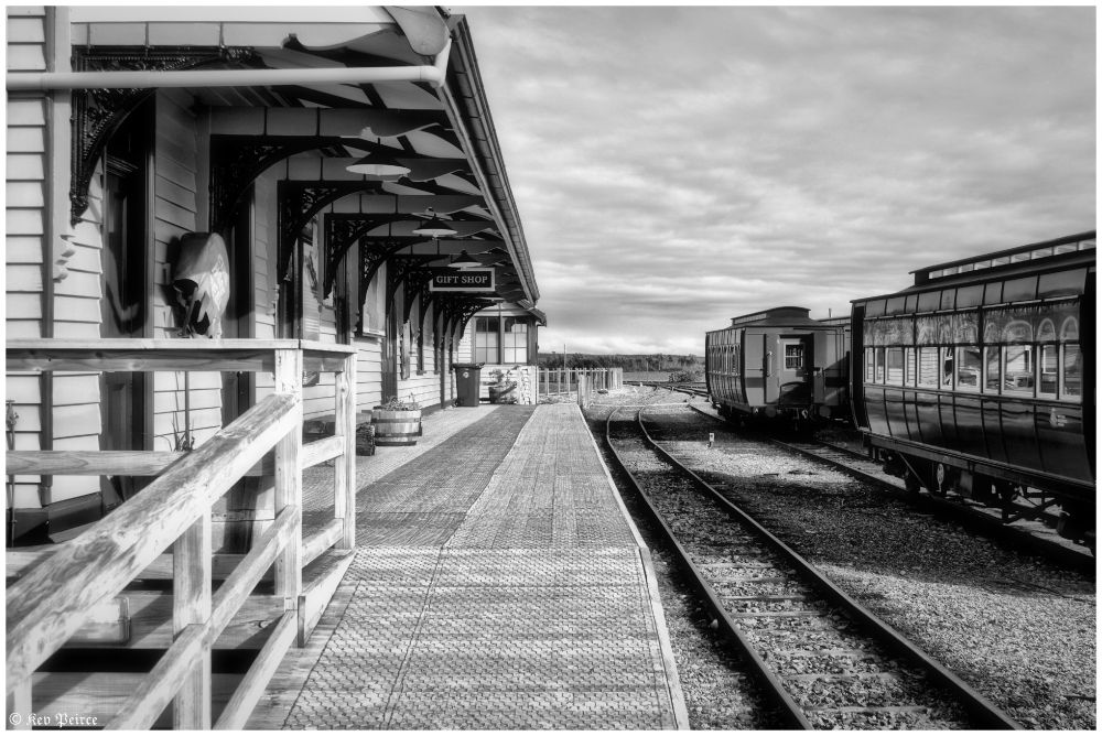 Black and white photo of a vintage railway station with timber platform, ornate ironwork, and historic train carriages.

A ‘Gift Shop’ sign hangs above the platform, evoking a nostalgic, early 20th-century atmosphere.