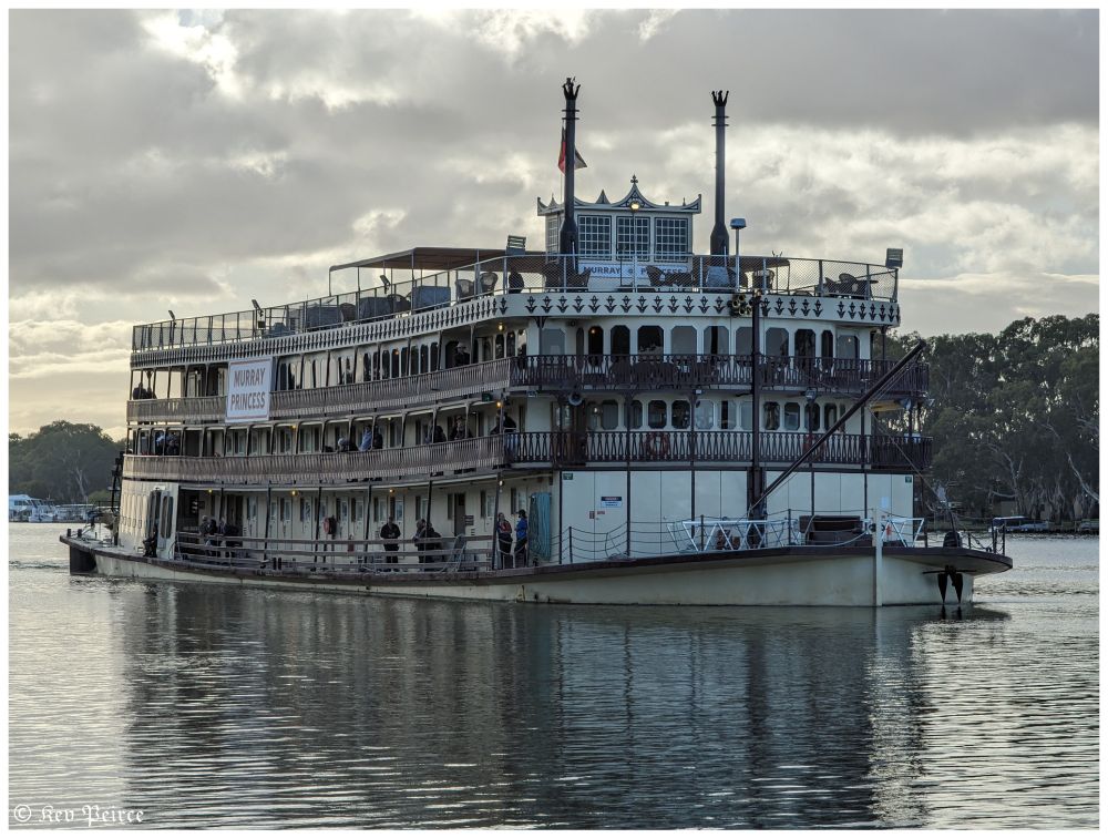 A grand paddle steamer named Murray Princess glides across a tranquil river under an overcast sky.

The multi-decked vessel features cream-colored railings, large windows, and visible passengers enjoying the journey. Lush trees line the riverbank in the distance, adding to the serene South Australian landscape.