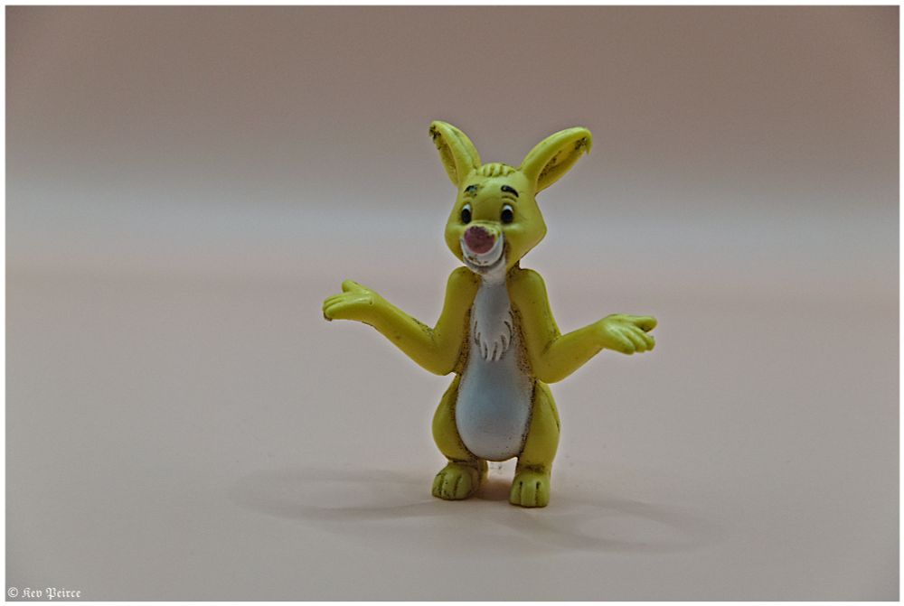 A small, yellow plastic toy rabbit figurine stands inside a lightbox. The rabbit has its arms outstretched, palms up, in a shrug-like gesture with a bewildered or confused expression on its face.