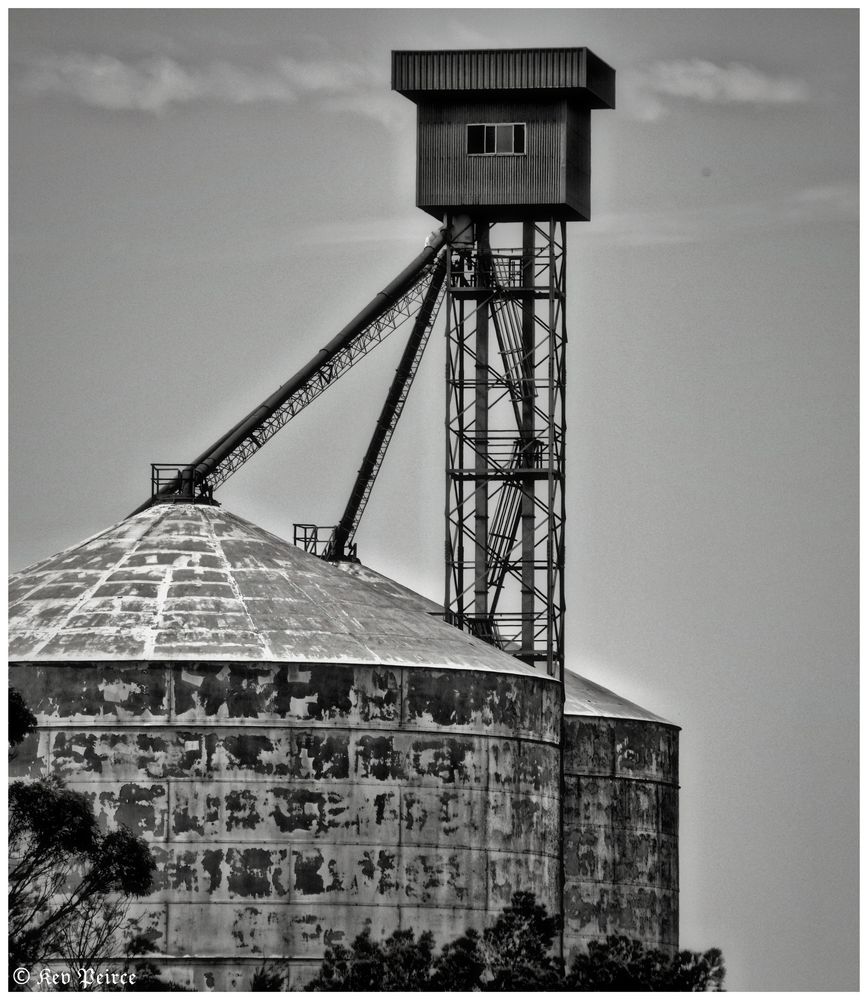 A black and white photograph showing two large, weathered grain silos in Kyancutta, South Australia. The one on the left is prominent in the foreground, with its heavily rusted, corrugated metal surface showing years of use.

A smaller, taller tower with a raised cabin at the top stands behind it, connected by an inclined conveyor belt structure. The sky is partially cloudy, and the base of the silos is partially obscured by trees.

The image is watermarked with the name "Kev Peirce" in the bottom-left corner.