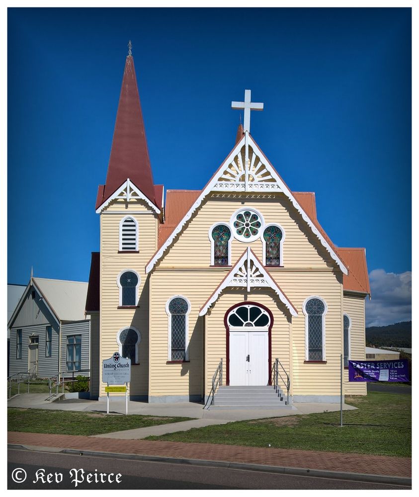 A historic Uniting Church stands beneath a clear blue sky, showcasing Gothic Revival architecture. Its tall steeple is topped with a cross, and the light yellow facade is accented by white trim and a red roof. 

Intricate woodwork and stained glass windows add character. A sign reads “Uniting Church,” and a lawn banner promotes “Easter Services.” 

The grounds are tidy, and the image is credited to Kev Peirce in the bottom left corner.