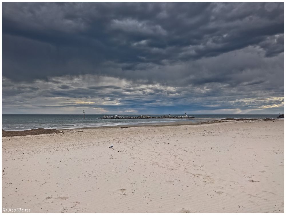 A moody beachscape under a brooding sky. Pale sand stretches toward a calm ocean, where gentle waves meet a rocky breakwater. 

Two navigational markers stand sentinel in the distance. Above, dark clouds churn, hinting at an approaching storm.

The scene balances serenity with tension, evoking quiet anticipation.