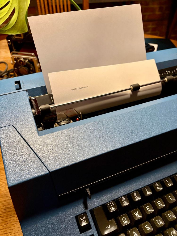 A photo of the top left corner of a beautifully blue IBM Selecteic II with a sheet of paper loaded in that says “Hello Mastodon”