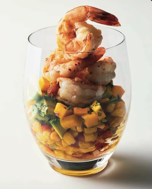 A glass filled with layers of colorful diced mango, avocado, and corn salad, topped with large cooked shrimp seasoned with spices and herbs.