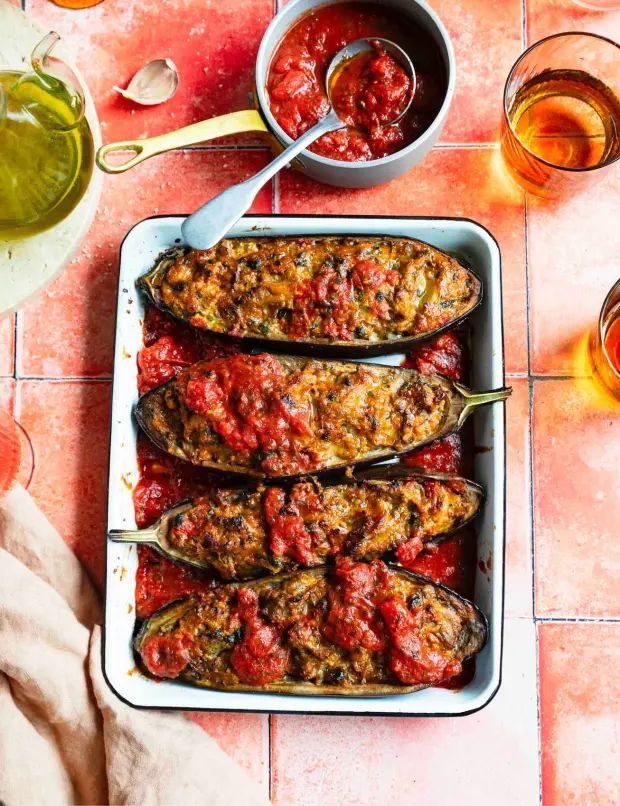 Four baked aubergine halves filled with a golden, savory stuffing and topped with rich tomato sauce sit in a white enamel baking dish on a rustic red tile surface. A small pot of tomato sauce, glasses of amber drink, olive oil, and a garlic clove surround the dish, creating a warm, inviting Mediterranean scene.