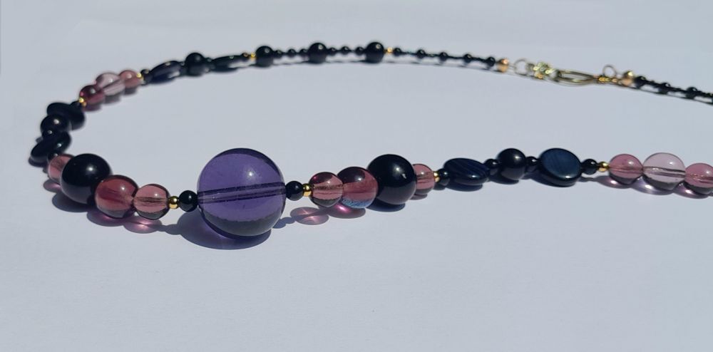 Necklace with round glass beads in shades of purple and black accented with small, gold beads, one larger purple glass bead at the centre