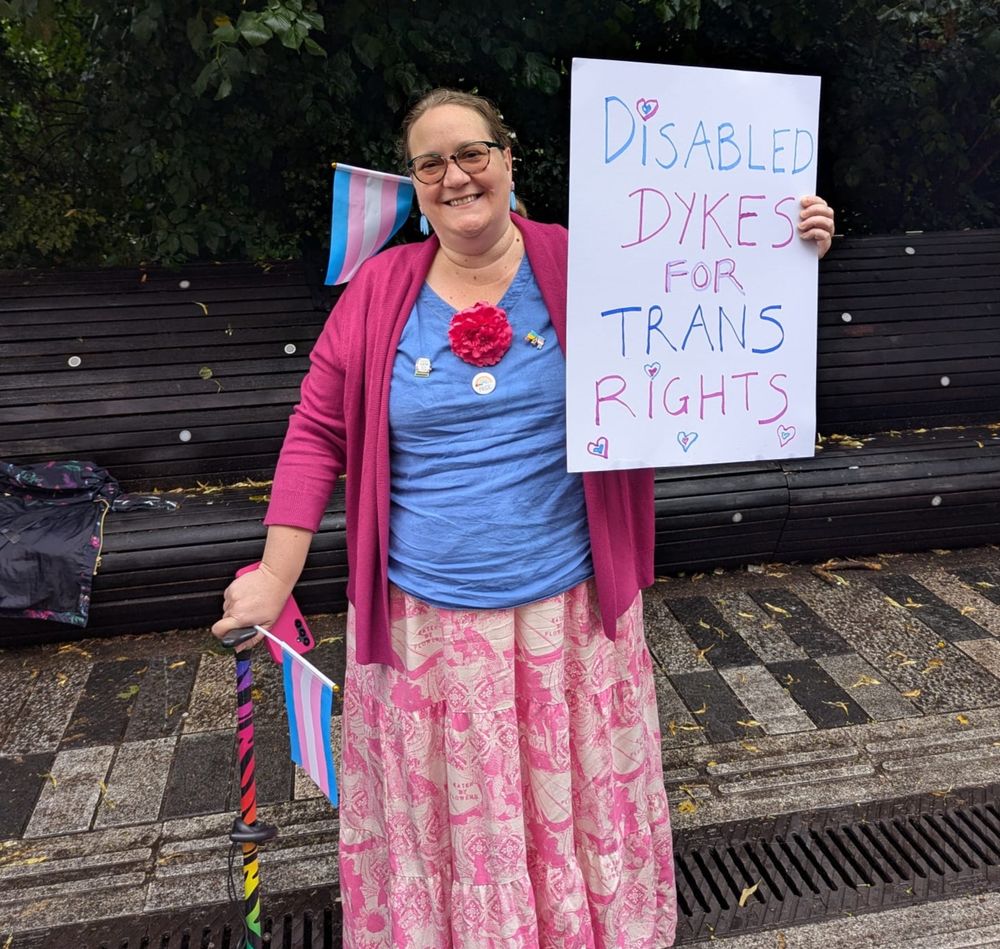 Person smiling at the camera, smiling, wearing colourful clothes and using a walking stick, holding a banner saying "Disabled dykes for trans rights"