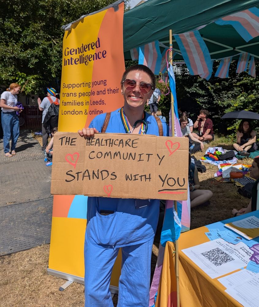 Person with short hair and sunglasses, wearing blue medical scrubs, smiling at the camera and holding a sign saying "The healthcare community stands with you"