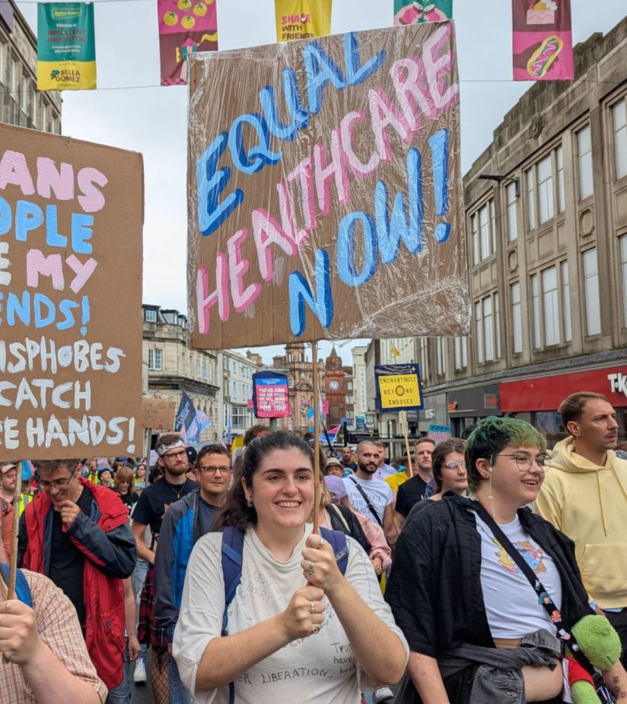 Photo of someone smiling, looking straight ahead and carrying a banner that reads "equal healthcare now!"