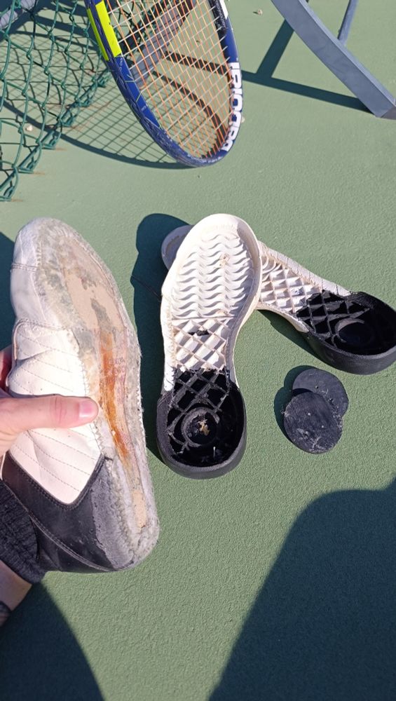 Une chaussure de tennis sans semelle, deux semelles décollées a côté. On voit une raquette de tennis au fonc