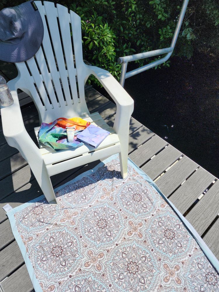 A patterned towel on a dock on a pond. A plastic Adirondack chair holds a sunhat, tie-dyed cotton shoulder bag, a bottle of sunscreen, and an ebook reader in a light purple case. 