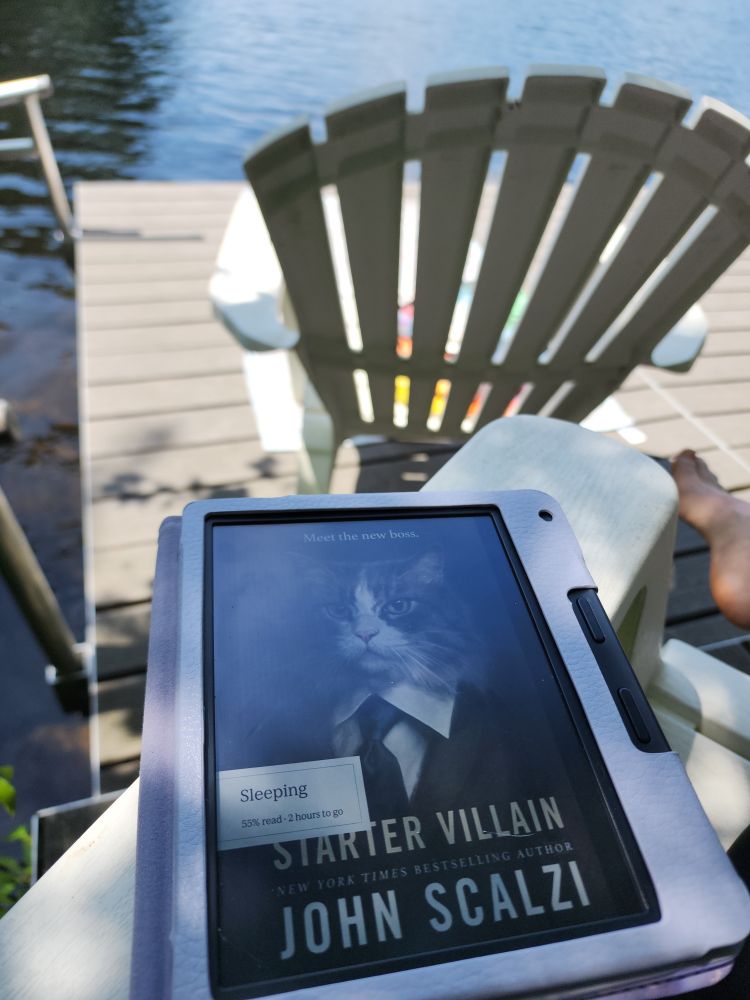 In the foreground, a Kobo Libra Colour, showing the cover of John Scalzi's "Starter Villain", on a plastic white Adirondack chair. In the background, a dock, with another chair and a towel, and a small lake/ pond of water beyond that. 