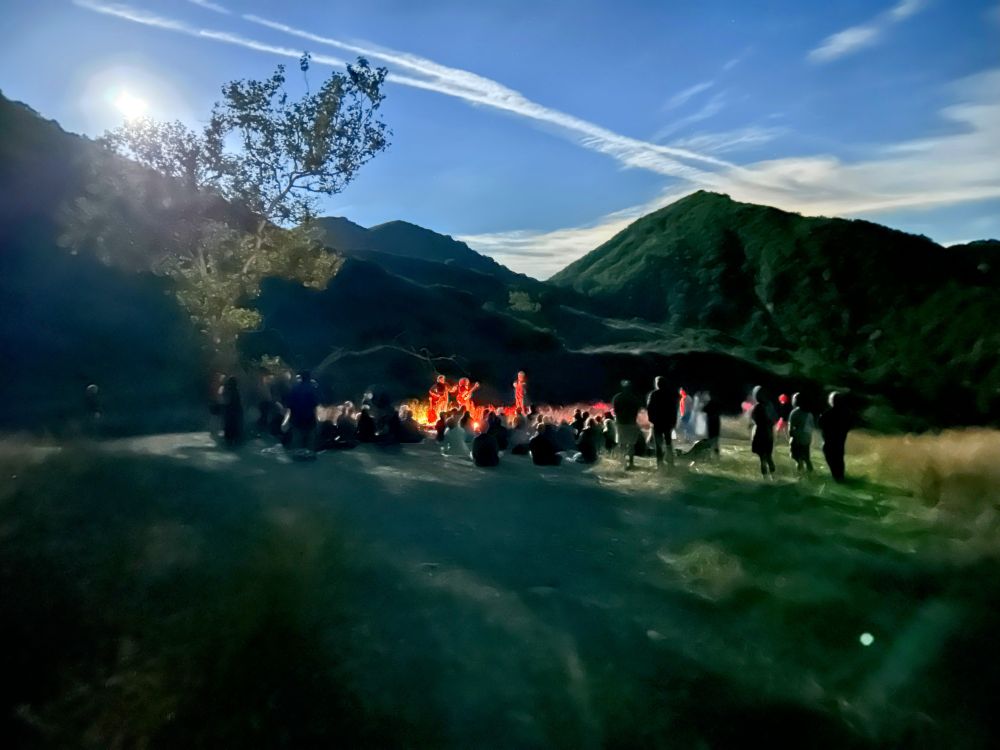 Moonrise behind an oak tree, a crowd gathered around two musicians who are lit in a warm glow. 