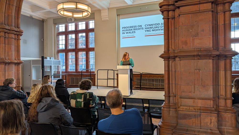 Sioned Williams is standing behind a lectern on a stage in front of rows of people who are seated. 