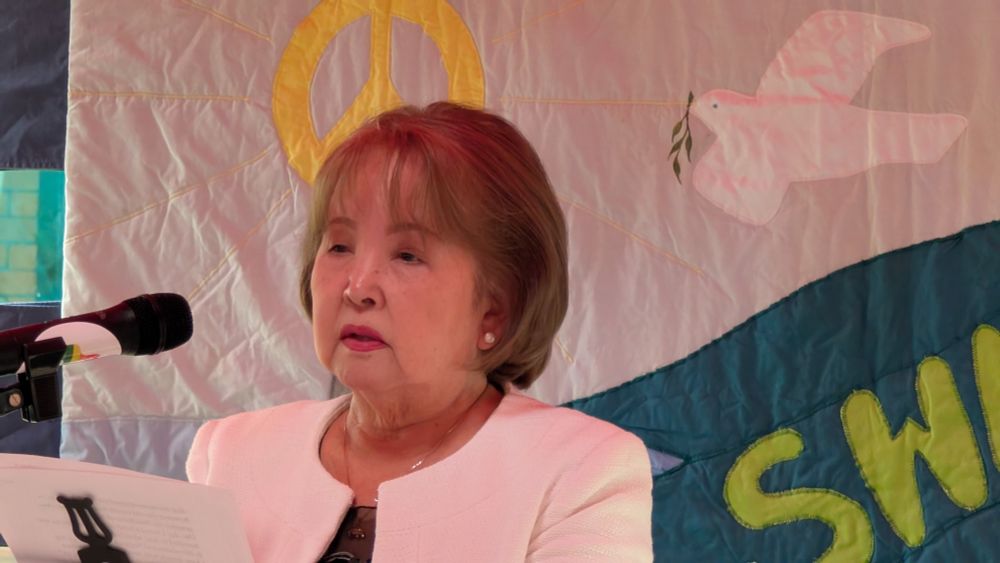 Kyoko Gibson speaking into a microphone in front of a CND banner.
