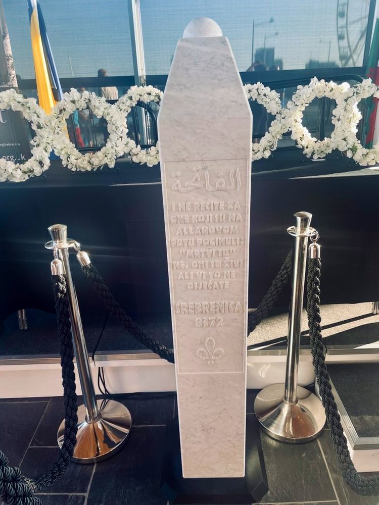 A white marble memorial stone to mark the Srebrenica Genocide standing on the floor of the Senedd with wreaths of white flowers set on a table behind it.