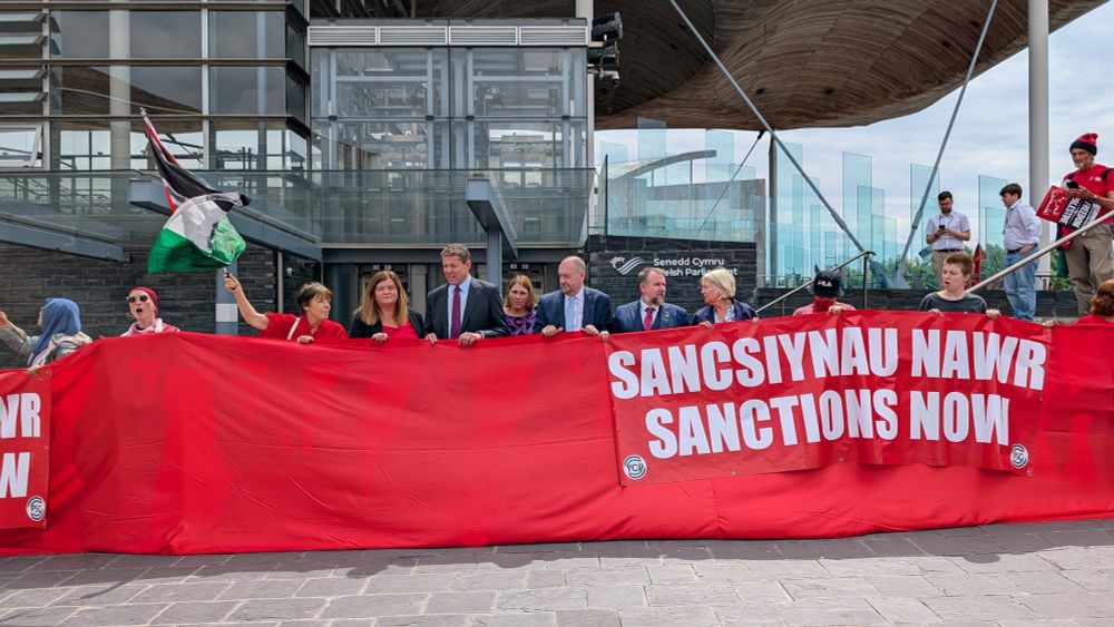 Sioned Williams MS stands with other members of the Plaid Cymru Senedd group at the red line protest. They are all behind the red “line” which is a long strip of material they are holding up that is covering from their feet up to their chests, so that you can only see their head and shoulder above the line. There is also a large banner that says “Sancsiynau Nawr / Sanctions Now” that is being held in front of the red line. They are in front of the Senedd. 