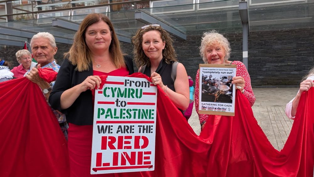 Sioned Williams MS stands with Sarah Rees from Oxfam Cymru and others holding up a long red “line” of material. Sioned and Sarah are also holding a sign that says “From Cymru to Palestine, we are the red line.” They are outside the Senedd. 