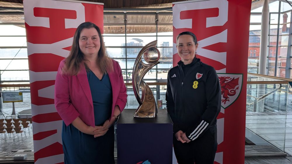 Sioned Williams MS stands with Rhian Wilkinson, either side of a table upon which sits the UEFA Women's EURO 2025 tournament trophy. Behind them are pop up banners with FAW logos and the word CYMRU in large letters running sideways on the banner, from top to bottom. They are inside the Senedd.