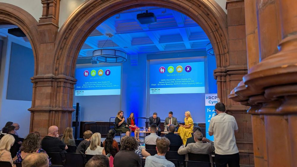 Sioned Williams is on a stage with 4 other members of a panel. Sioned is holding a microphone. They are at an event inside the main hall of the Pierhead Building, and the photo is taken from behind the audience. A large archway spans over their heads, and the stage can be seen through the archway. 