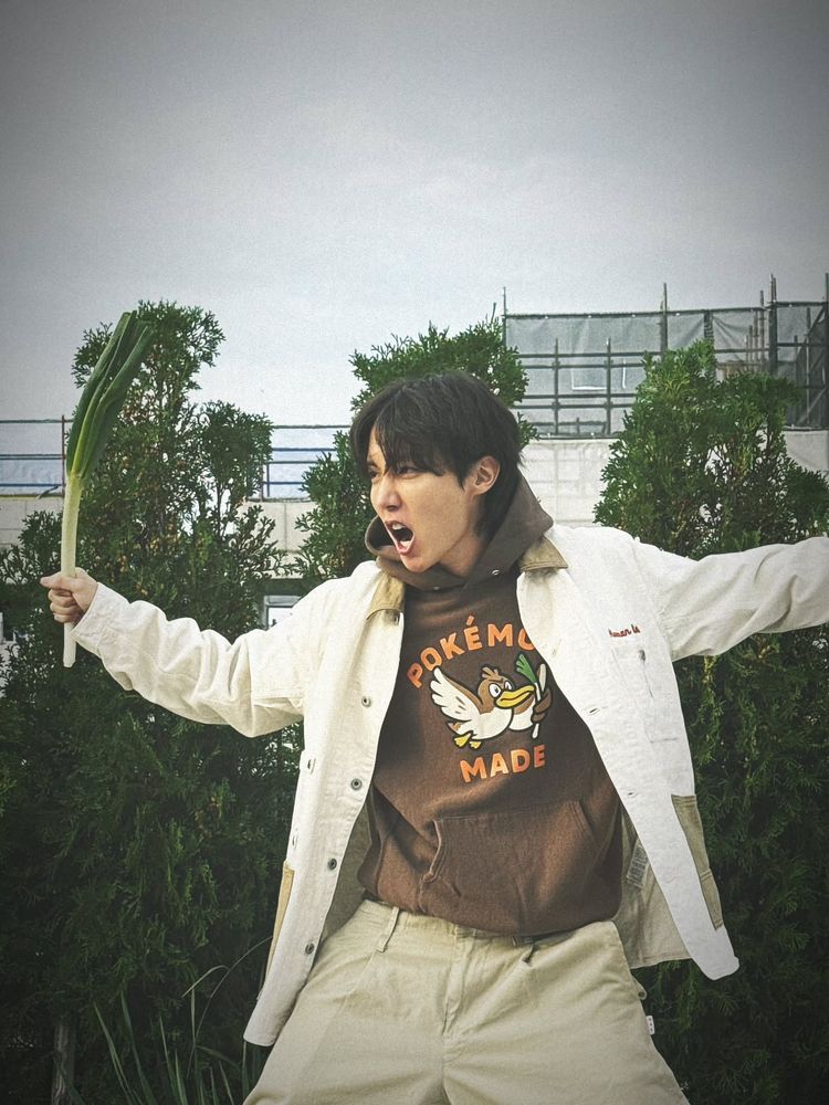 jhope do bts, com uma blusa de pokémon marrom, uma calça bege e um casaco branco. ele segura um alho poró como se fosse uma espada. 