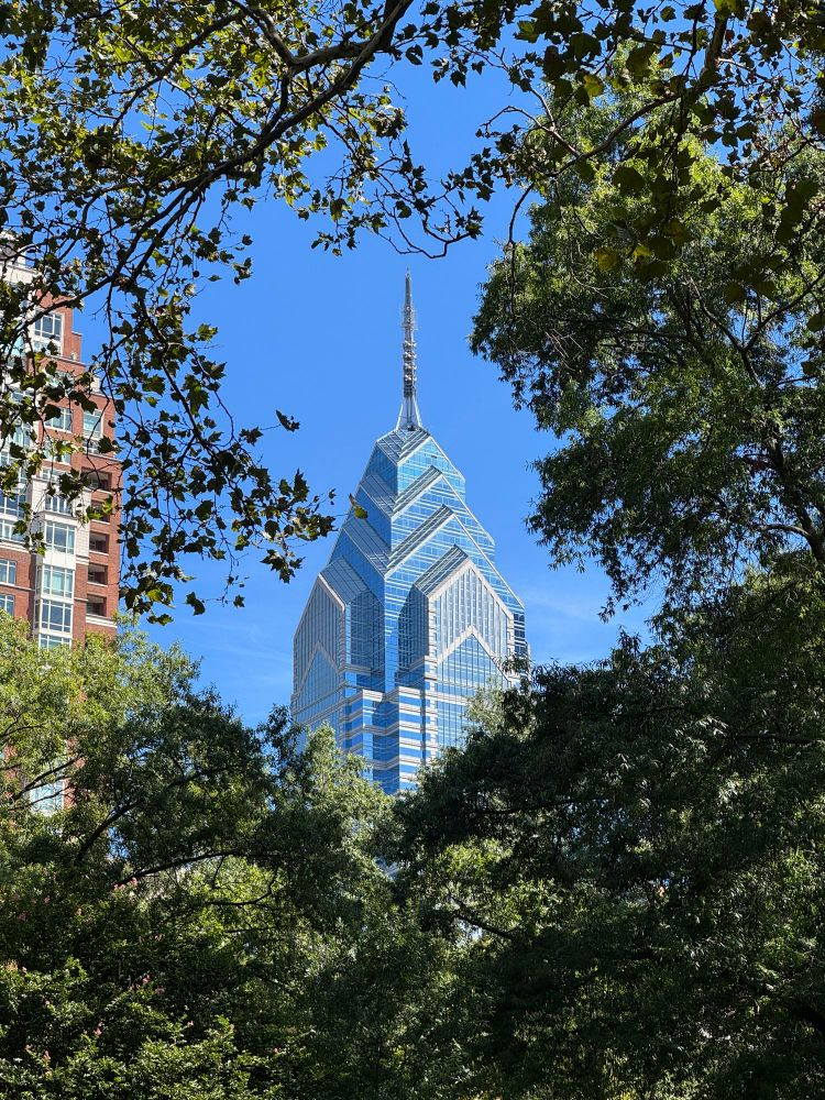 Blue glass skyscraper with stepped pyramidal spire rises behind the green tree canopy in Rittenhouse Square Park Philadelphia