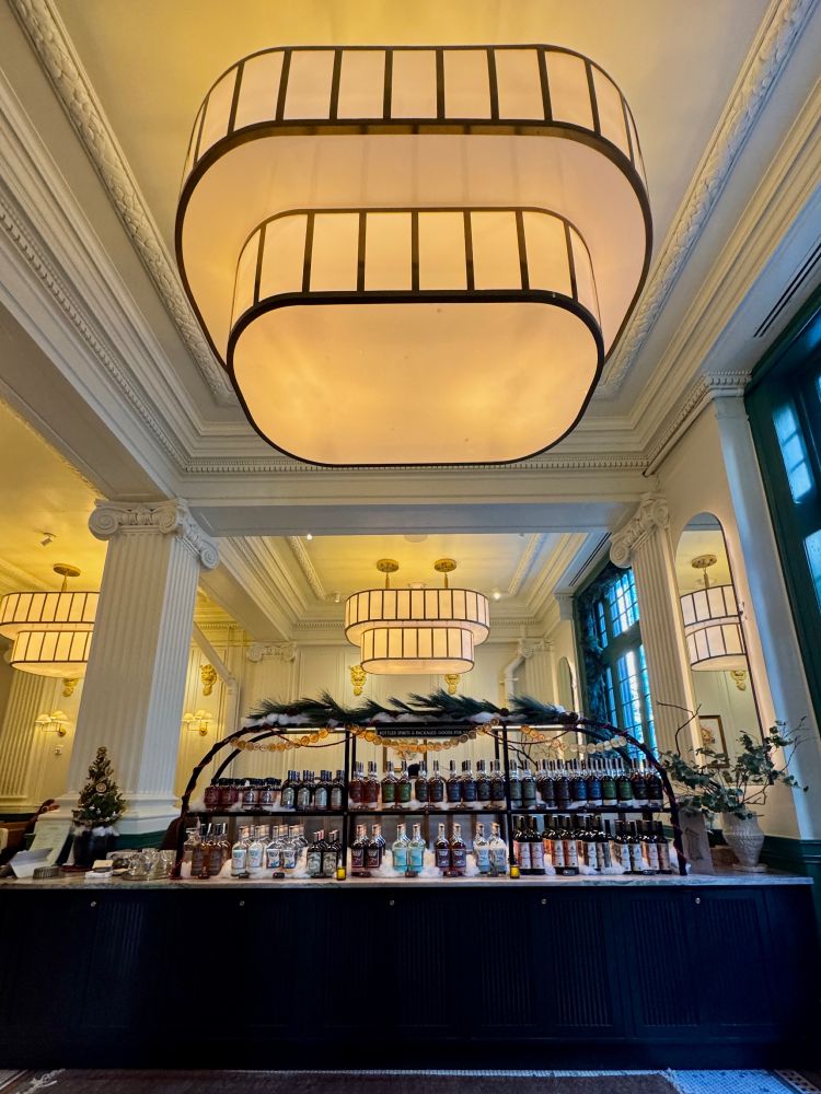 Tall ceiling in a dramatic dining space with elaborate pilasters, moldings, and light fixtures; a rack of craft alcohol varieties sits below a festive garland