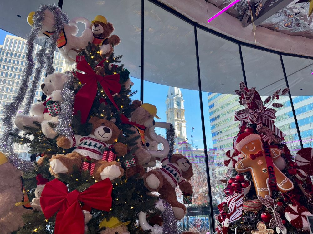 Two decorated Christmas/holiday trees against a wall of windows frame a view of the tower of Philadelphia’s City Hall. Tree on left has teddy bears in sweaters, on right tree has peppermints and a large gingerbread man