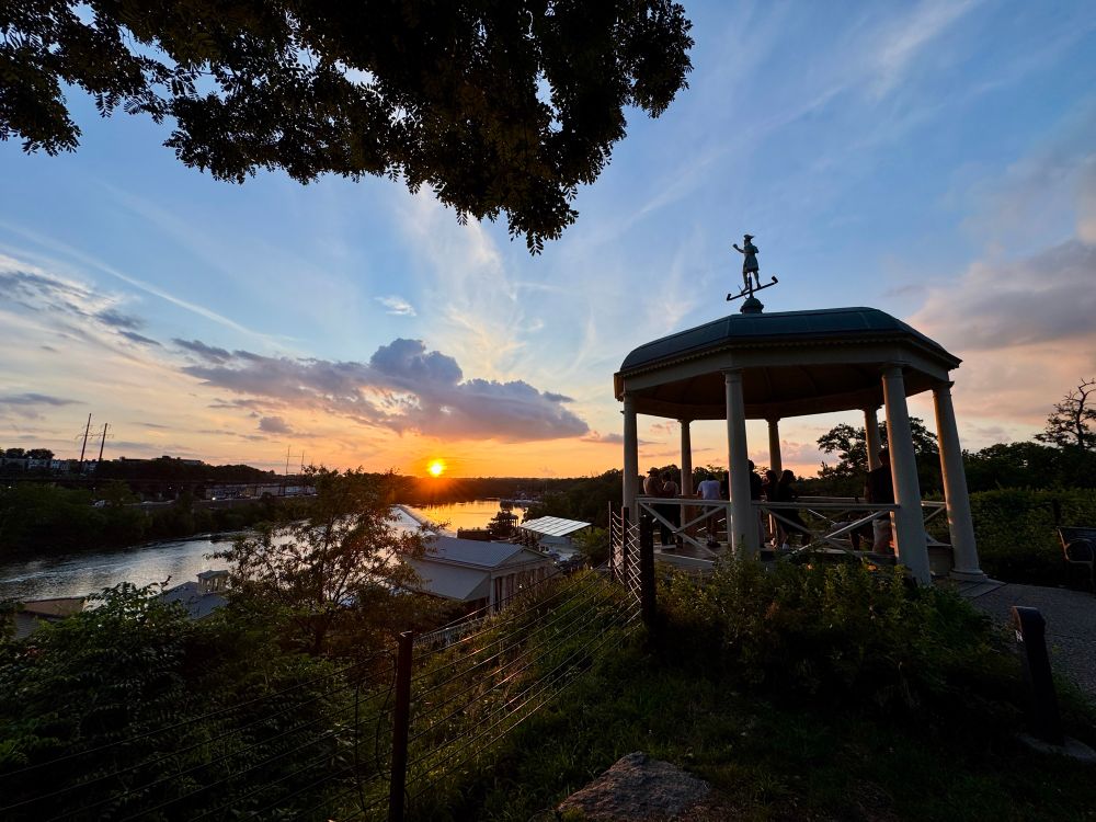 A white gazebo perched on a cliff is kissed by an orange glow, above a glimmering river at sunset