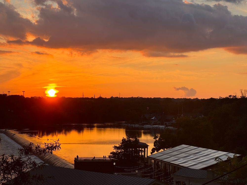 the orange glow of the setting sun sets the skies ablaze over west philadelphia, reflecting on the glassy water below