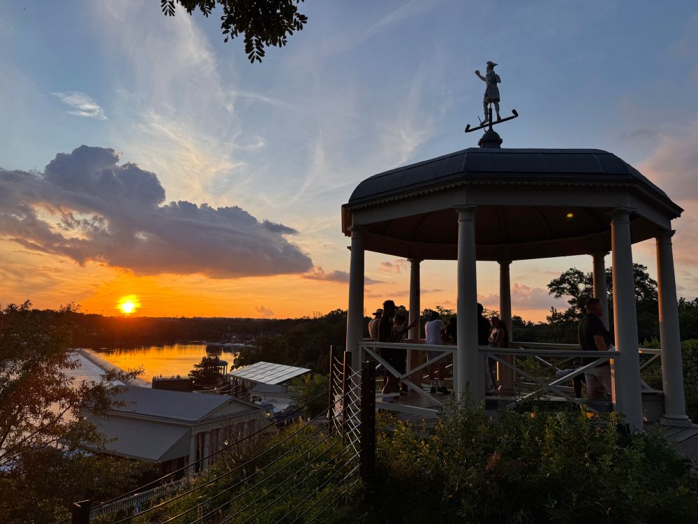 A figure of Roman god Mercury crowns an octagonal wooden pavilion full of onlookers high above the Fairmount Waterworks historical site on the Schuylkill River during an epic sunset moment 
