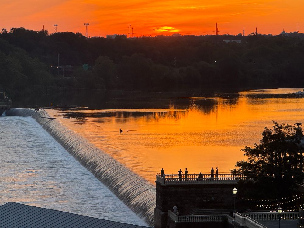 people in silhouette on a pier overlooking a dam at sunset, the still water of the glassy upstream pool reflecting the vermilion skies 