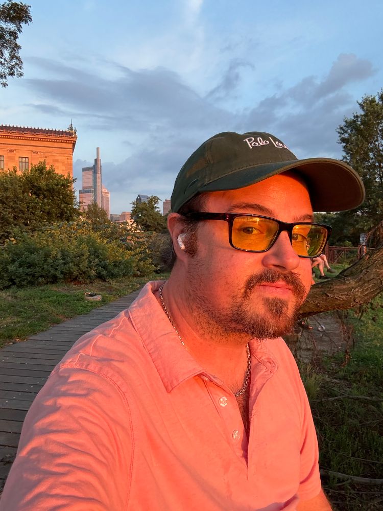 your photographer in a pink polo, glasses, and green cap, smiling at camera. philadelphia museum of art is behind on left, comcast center four seasons hotel tower of philly skyline in distance 