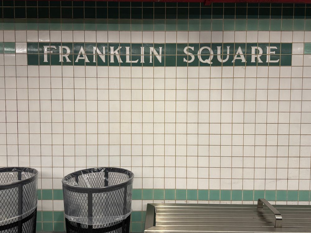 vintage green and white tiles on train platform reading Franklin Square, two trash cans and a long stainless steel bench 