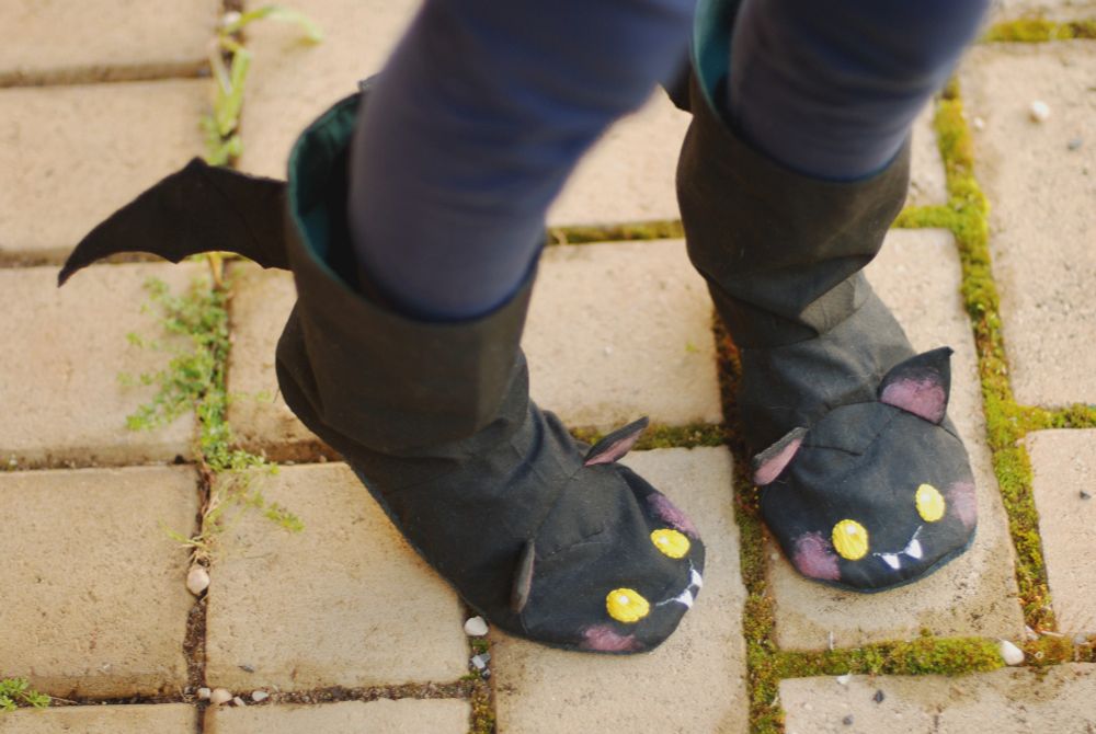 A pair of slippers that are also bats, sewn in black cotton fabric with bat wings, green lining, with embroidered yellow eyes, white fangs, and other details applied with fabric paint.