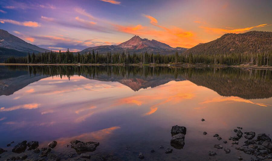 A vivid Oregon sunset over Sparks Lake. The still water perfectly reflects the glowing sky and the distant peaks of the Three Sisters, surrounded by pine trees and soft evening light.