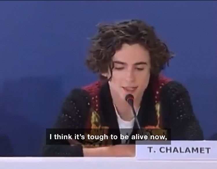Timothee Chalamet, a thin young white man in a navy, burgundy, and yellow sweater, sits behind a mic at a table and says, “I think it’s tough to be alive now, I think societal collapse is in the air, it smells like it.”