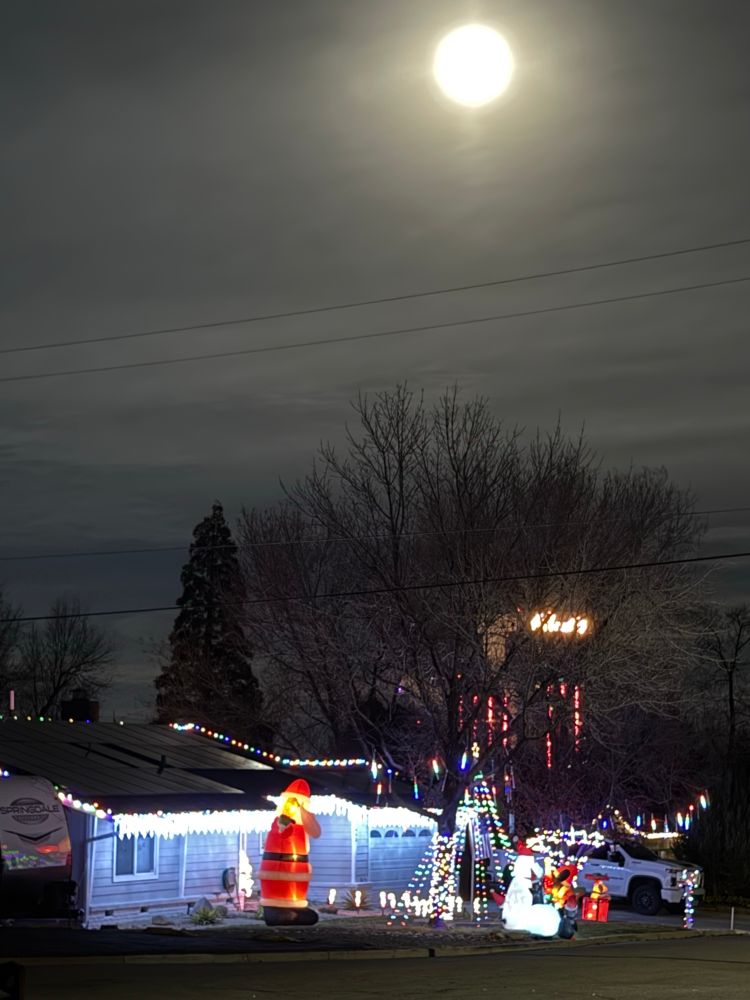 A bright full moon shines through a hazy layer of clouds above trees and a house with several elaborate holiday lawn decorations.  A hotel-casino resort can be seen behind the trees.