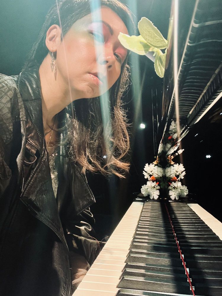 Une pianiste en veste de cuir noir devant piano à queue noir et en mode autoportrait, quelques fleurs décorent le piano 