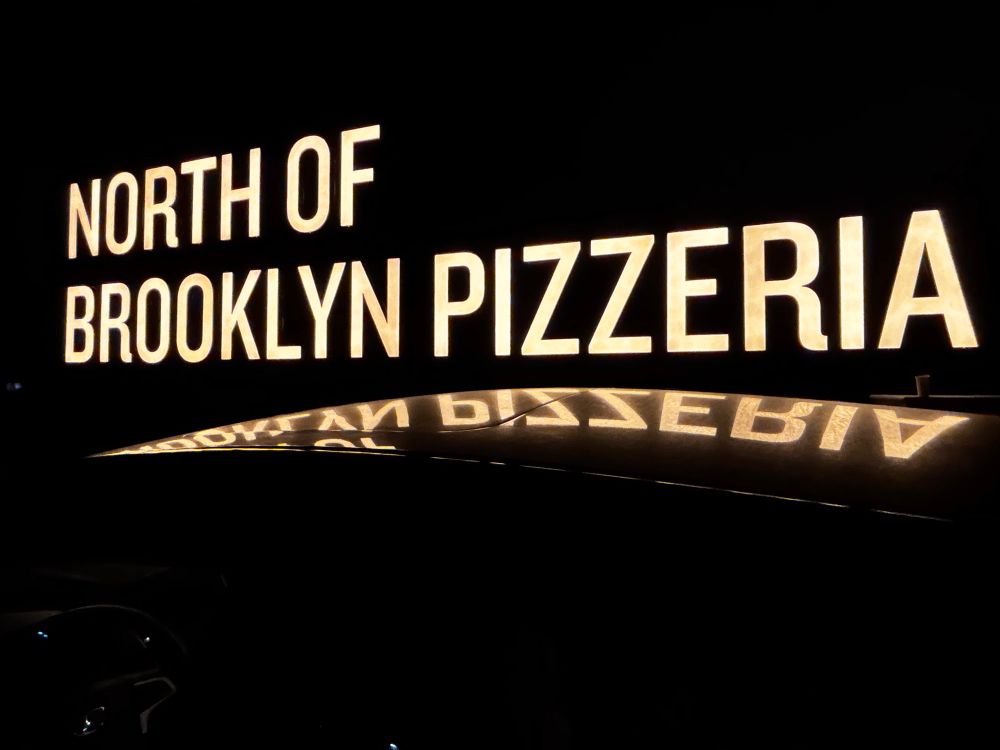 North of Brooklyn Pizzeria signage lit up at night and reflecting off car roof 