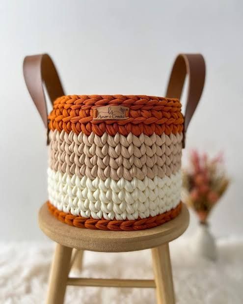 Um cachepô feito com fio de malha nas cores amarelo clarinho, marrom claro e terra. Dos lados, tem uma alça em couro 