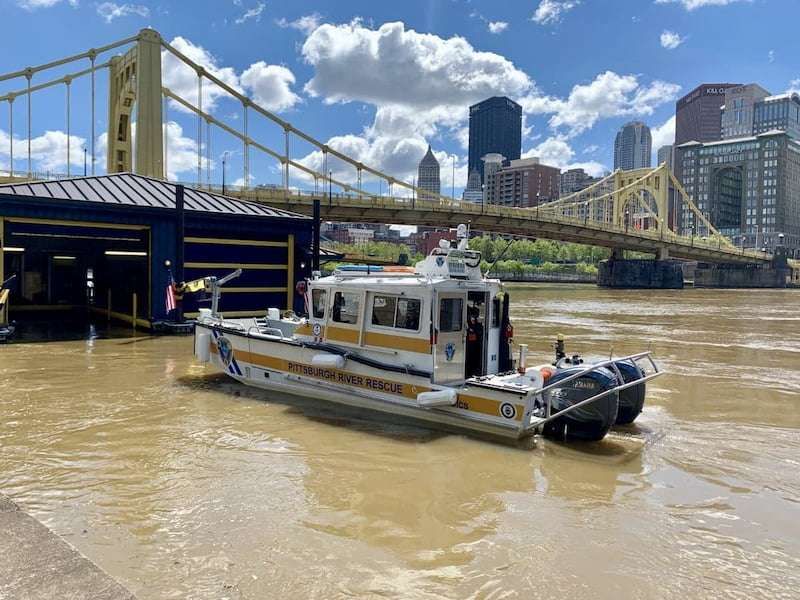 A Pittsburgh River Rescue boat on the water. Boat is white with two horizontal yellow stripes. The boat is headed away from the camera, towards a bridge.